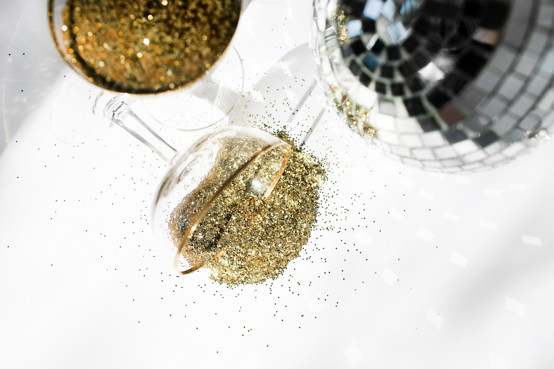A silver disco ball and a champagne glass laying in glitter on a white surface | S'Berry Boutique